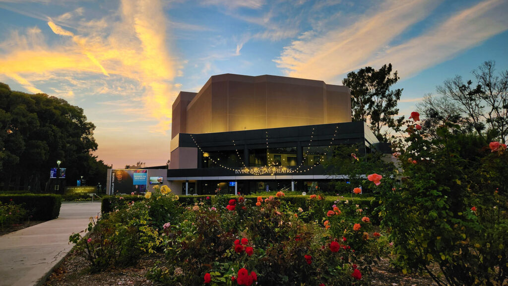 Exterior of the Irvine Barclay Theatre at dusk, with warm sunset clouds overhead, string lights strung across the entrance, and a rose garden in the foreground.