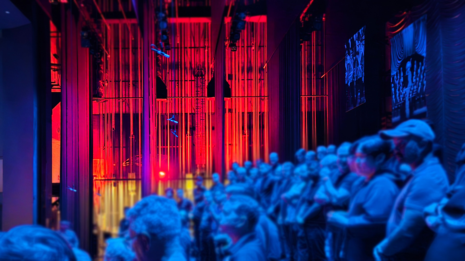 Backstage view looking out toward a stage bathed in red light, with rigging and rails overhead. A group of choir members in blue stage lighting stands gathered in the wings, waiting to perform.